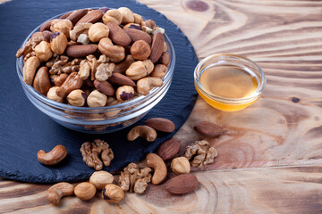 Assorted nuts in glass bowl with honey bowl. Mixed nuts on wooden table. Black stone plate on wooden background.