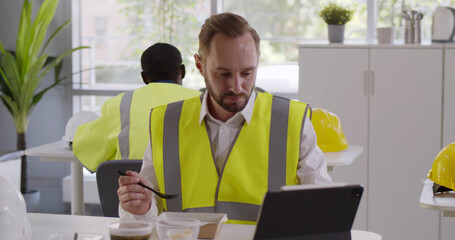 Construction company worker in reflective vest having lunch and using digital tablet in canteen