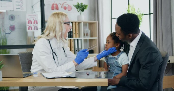 Healthcare and medicine concept where charming positive kind skilled blond female doctor checking girl's throat with spatula during general inspection while girl visit clinic with caring balck-skinned