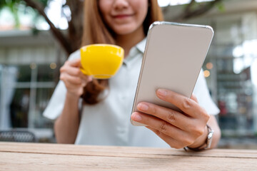 Closeup image of a young asian woman holding and using mobile phone while drinking coffee