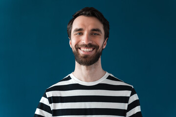 Young, smiling, bearded man on a blue background. The man in the striped T-shirt is smiling sincerely