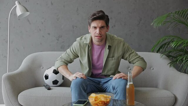 Young fun man fan in green clothes t-shirt cheer up support football team sit on grey sofa with soccer ball bottle beer ale eat chips rest watch tv indoors room gray color wall. Sport leisure concept
