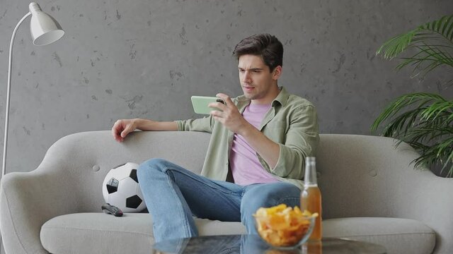 Young fun happy man fan in green clothes t-shirt cheer up support sport football team sit on grey sofa with soccer ball bottle beer eat chips wait watch tv on mobile cell phone indoors room gray wall