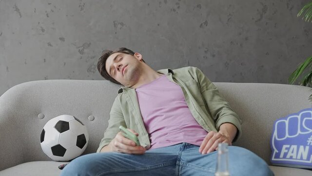 Young tired man fan in green clothes t-shirt cheer up support sport football team sit on grey sofa with soccer ball bottle beer eat chips wake up watch tv hold money mobile cell phone win indoor room