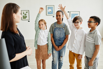 Diverse group of children raising hands while listening to female expert in modern art gallery