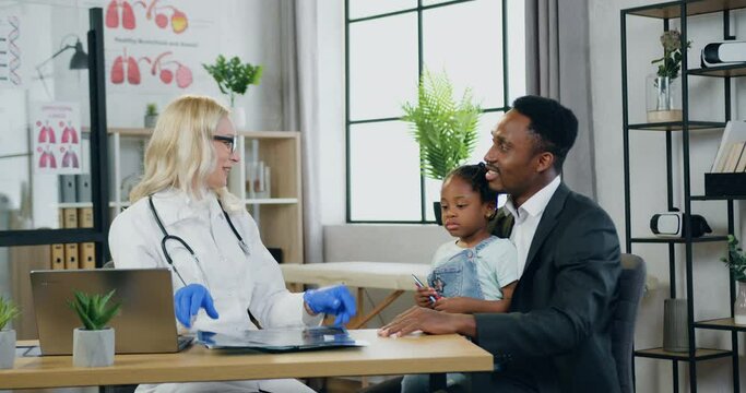 Pretty smiling professional blond woman-doctor explaining x-ray results of small girl's lungs to girl's father during their scheduled visit to medical center,medicine concept