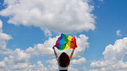 Homosexsual, lesbian, woman holding a rainbow LGBT gender identity flag on sky background with clouds on a sunny day and celebrating a gay parade, Bisexuality Day or National Coming Out Day
