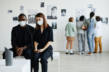Minimal portrait of mixed-race couple in art gallery wearing masks and listening to audio guide,...