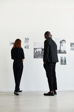 Minimal back view at two people wearing black while looking at photographs in modern art gallery