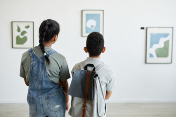 Back view at two schoolchildren visiting art gallery and looking at modern abstract paintings, copy...
