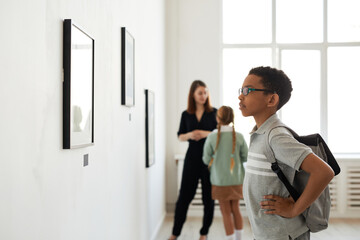 Side view portrait of young African-American schoolboy looking at paintings in modern art gallery, copy space