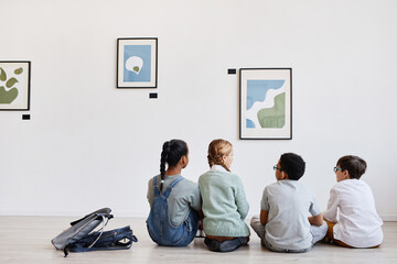 Back view at diverse group of children sitting on floor in modern art gallery and discussing...