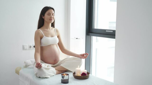 Pregnant woman relax doing yoga, sitting in lotus position in spa cabinet alone with candles, with eyes closed, keep calm. Health lifestyle pregnancy beauty, interior. Mom-to-be keep hands together