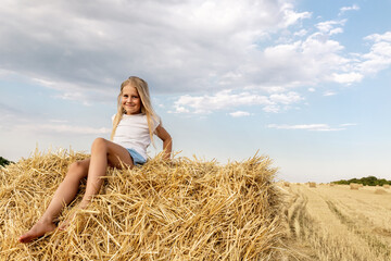Naklejka na ściany i meble Portrait of cute little blond beautiful adorable cheerful caucasian kid girl enjoy sitting on hay stack or bale on harvested wheat field warm summer evening. Scenic rural country landscape background