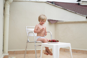 White child eats strawberries in summer sitting on the terrace on the background of the roofs