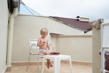 Boy eats strawberries in summer sitting on the terrace on the background of the roofs