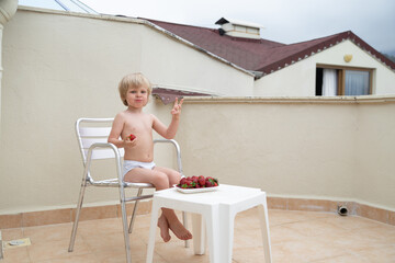 Caucasian child eats strawberries in summer sitting on the terrace on the background of the roofs