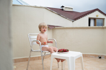 Cute child eats strawberries in summer sitting on the terrace on the background of the roofs