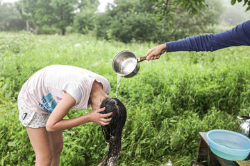 A teenage girl in a light T-shirt and shorts washes her hair from a bucket in nature, her face is not visible, shooting from the side, long dark hair, in daylight