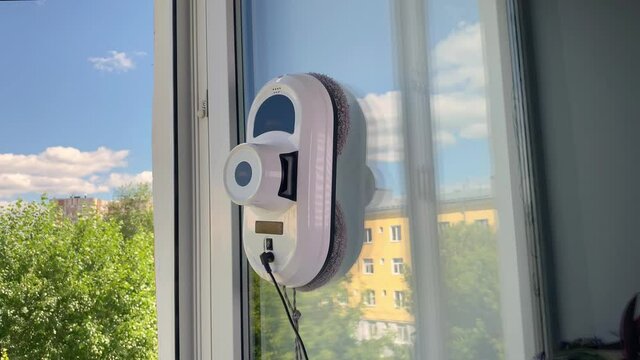 Domestic vacuum cleaner robot washing glass window in room. Blue sky and yellow building outside. Robot assistant cleaning windows