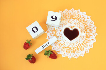 Calendar for July 9 : cubes with the numbers 0 and 9, the name of the month of July in English, a cup of tea in the shape of a heart on a white openwork napkin, strawberries on a yellow background