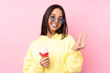 Young brunette girl holding a cornet ice cream over isolated pink background smiling and showing...