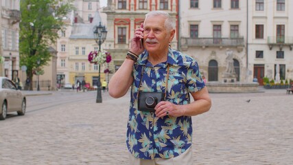 Fototapeta premium Senior grandfather enjoying smartphone conversation and traveling. Elderly old tourist man in stylish clothes talking on mobile phone while walking on city street. Mature people life after retirement