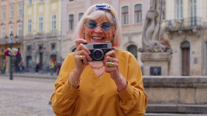 Portrait of senior woman tourist taking pictures with photo camera, smiling using retro device in summer city center of Lviv, Ukraine. Active life after retirement. Photography, travelling, vacation.