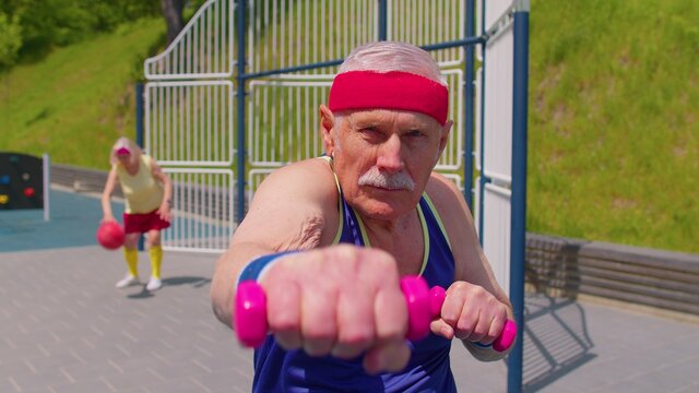 Active senior handsome man grandfather doing sport training boxing fitness aerobics cardio exercising with dumbbells on basketball playground. Healthy grandmother playing ball basketball on background