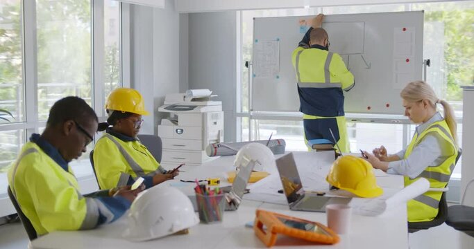 Diverse constructors sitting at desk and using smartphone with colleague writing on whiteboard