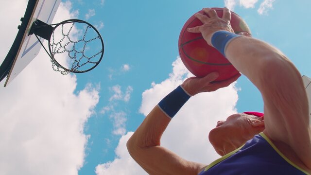 Upward View Of Focused Senior Player Man Playing Basketball Game, Successfully Hits Goal. Active Sportsman Grandfather Trains Throwing Ball Into Basket Ring On Sport Playground Yard