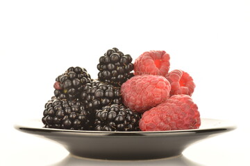 Ripe fresh organic blackberries and red raspberries on a black ceramic saucer, close-up, isolated on white.