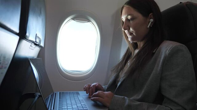 Brunette in suit with wireless earphone in ear works at laptop while sitting on airplane.