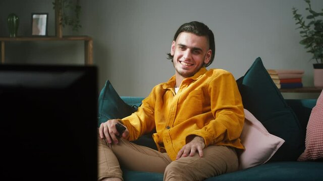 Young man is sitting on a sofa and watching entertainment program on television. The guy watches funny film on TV and laughing, spending free time in living room at home. 