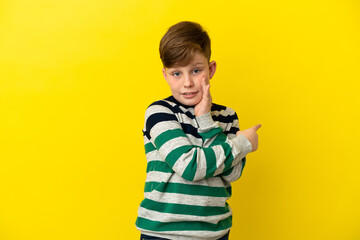 Little redhead boy isolated on yellow background pointing to the side to present a product and whispering something