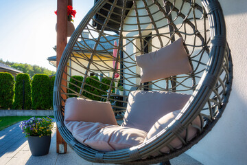 Modern hanging rattan chair with gray pillow and flowers on the summer garden terrace
