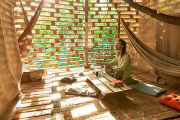 Obraz premium Zoomed out view of young longhaired man meditates in the wooden terrace 