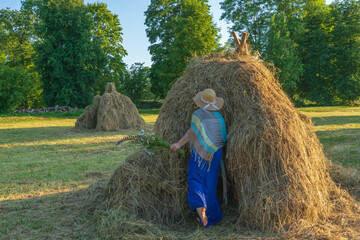 A woman in a straw cap with a bouquet of field flowers near a pile of hay © Ieva