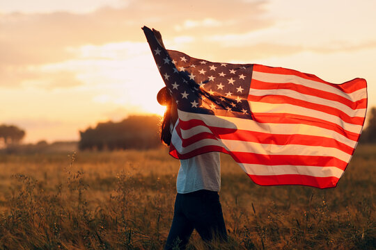 Woman farmer in wheat field waving american flag on sunset.