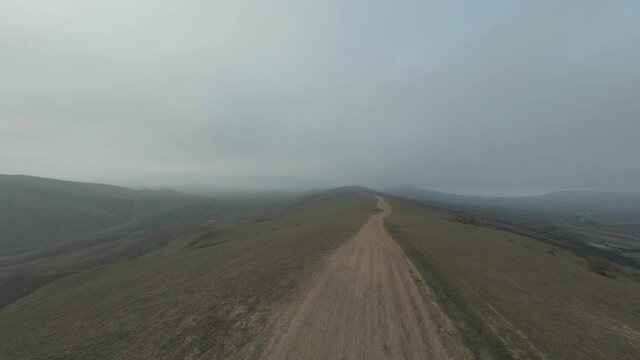 Buggy vehicles drive along brown ground road running on hill ridge between fields against mountains on horizon. First person fpv drone view