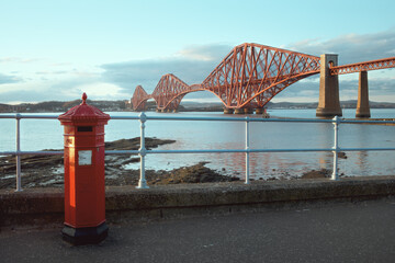 A red vintage British post box in front of the iconic Forth Rail Bridge in South Queensferry,...