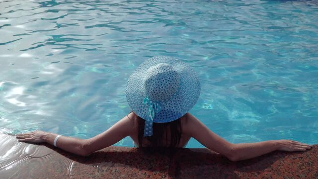Back of a woman in blue hat leaning on pool border inside swimming pool covered with Mozaic tile, template copy space, elevated view