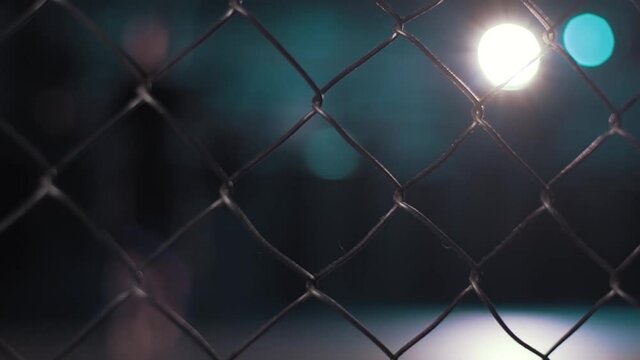 Close up of chain link fence on dark blurred background with shining lamp. Video. Metal lattice inside the building, grunge style.