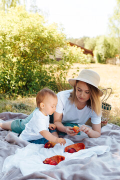 Young mother and little son on picnic in nature. Beautiful mom in hat feeding fruit to her baby. Happy family having a rest in the summer, outdoors. Vertical photo