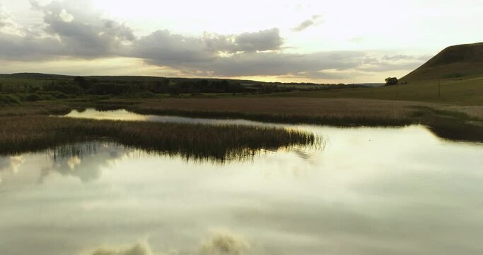 Source of the river with beauty calm swampy backwater with reeds and thickets and bald hills background. Aerial above drone wide view at summer evening sky.