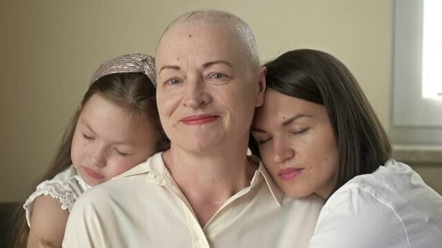 An adult daughter and little granddaughter hug a sad elderly woman who has gone bald after chemotherapy. Love, care and support in the family of a cancer patient.