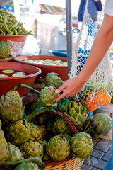 Plant based diet concept. Young woman with reusable eco friendly net bag picking fresh artichokes on farmers market. Conscious shopping for organic local vegetables. Close up, copy space, background.
