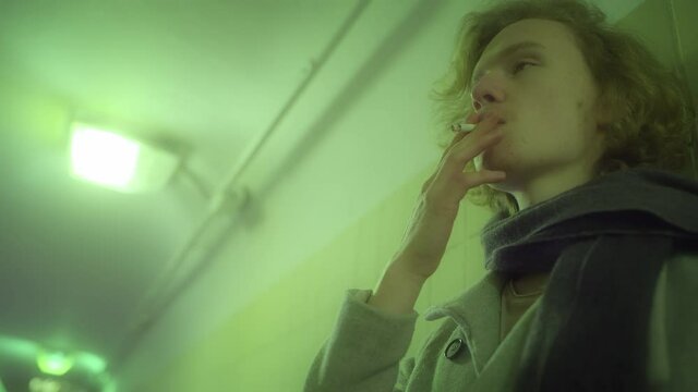 An attractive caucasian man blond with long hair in a coat stands in the underpass, smokes a cigarette and looks at the camera