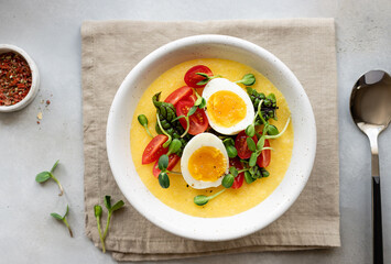 polenta served with fresh vegetables, microgreens and eggs in white bowl on linen napkin. Healthy breakfast or lunch. flat lay