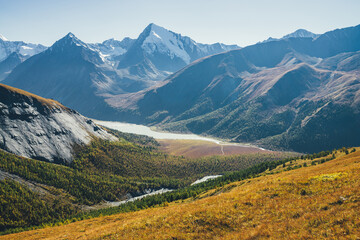 Obraz premium Wonderful alpine landscape with mountain lake and mountain river in valley with forest in autumn colors on background of snowy mountains silhouettes under blue sky. Beautiful mountain valley in autumn